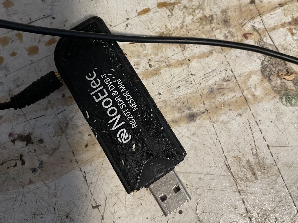 A well-used NooElec NESDR Mini R820T SDR USB dongle with its antenna cable on a paint-stained workbench