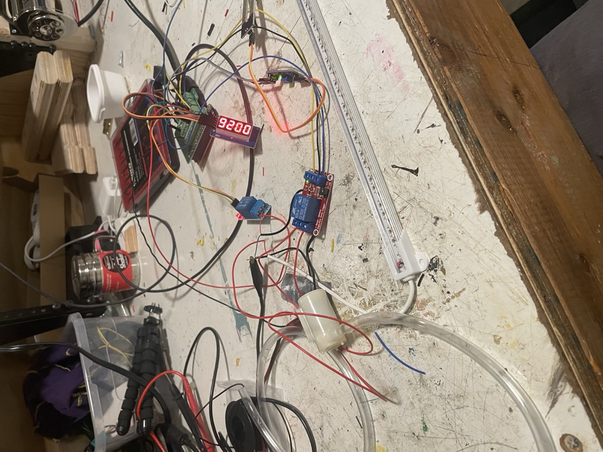 A workbench covered in prototyping parts: a pump, relay modules, a soil moisture sensor, an LED grow light strip, and a seven-segment display
